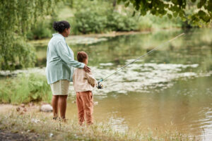 Back view of mother and daughter fishing together by lake in beautiful family bonding moment, copy space