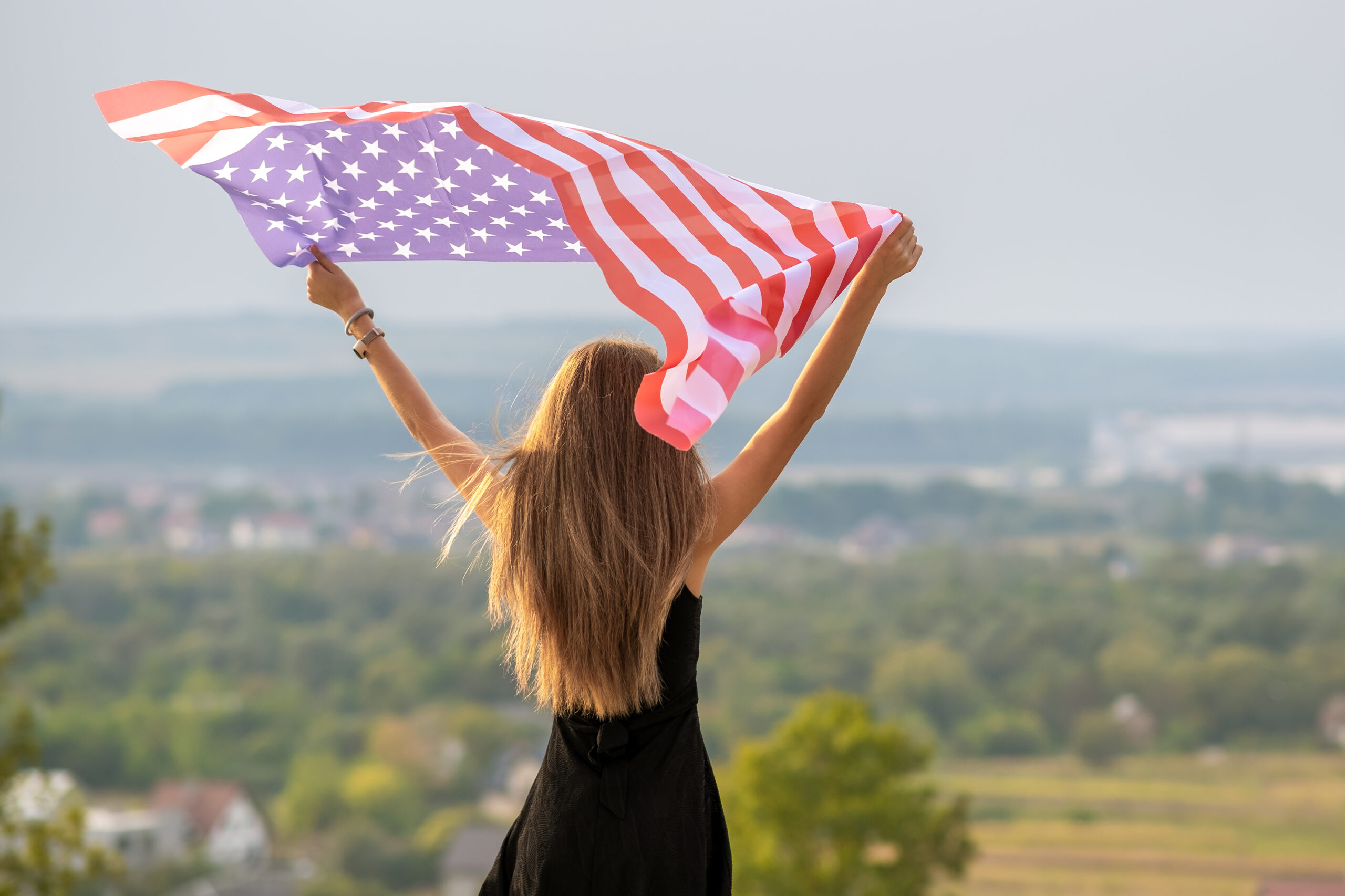 Young happy american woman with long hair raising up waving on wind USA national flag in her hands relaxing outdoors enjoying warm summer day.