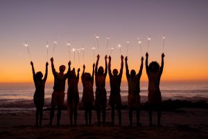 A multi-ethnic group of friends enjoying their time together on a beach during sunset, standing in a row, holding sparklers, raising their arms